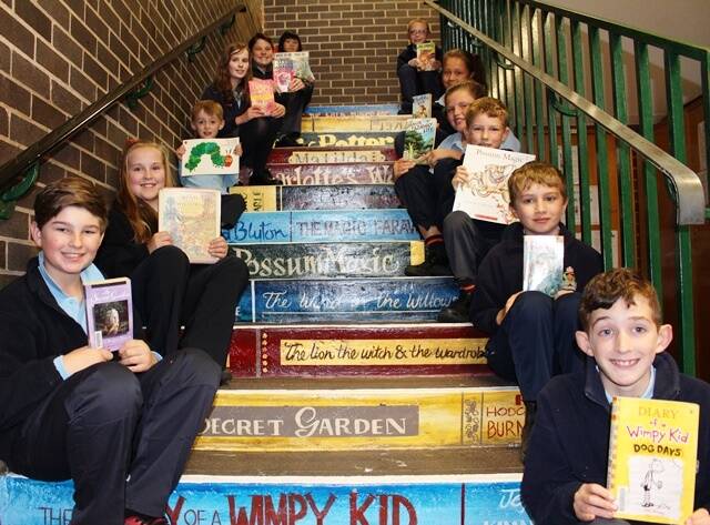 Pictured on the amazing stairs leading up to the school library are students from Kindergarten through to Year 6 with their favourite books.
The stairs leading to the library and Year 6 room have recently had an amazing transformation from dull and grey to bright and wonderful. As the stairs lead to our library we thought it would be a great idea to turn the stair risers into spines of some of our favourite books. There are classics like The Wind in the Willows and The Lion, the Witch and the Wardrobe and some more recent favourites like Diary of a Wimpy Kid and Harry Potter. It has totally changed the look and the feel of the area thanks to the wonderful artistic skills of Cullen Ball, Lachlan and Cooper Tetley’s uncle. The challenge now is to see who can read all the titles painted on the stairs, how many have you read?